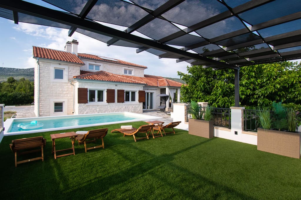 The villa, the pool and sunbeds on the lawn, seen from under the pergola