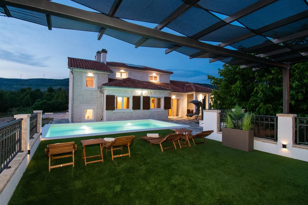 The villa and the lit pool at dusk, seen from under the pergola